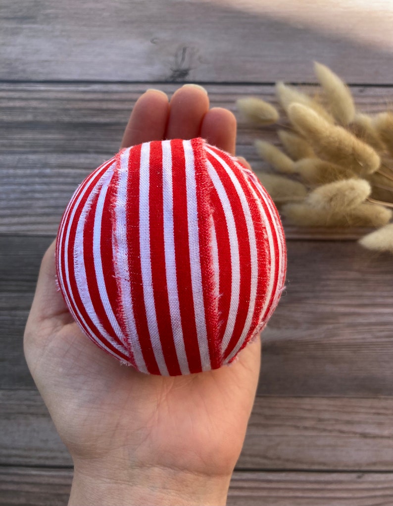 Puede incluir: Una bola de adorno de tela a rayas rojas y blancas. La bola est&aacute; hecha a mano y tiene una superficie ligeramente irregular.