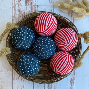 Puede incluir: Seis bolas de tela en una cesta de mimbre. Tres bolas son de rayas rojas y blancas y tres bolas son azules con estrellas blancas.