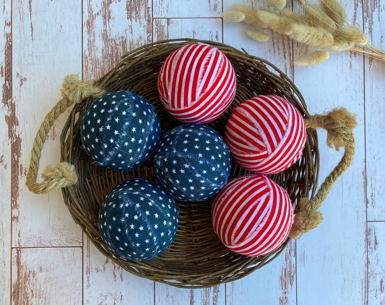 Puede incluir: Seis bolas de tela en una cesta de mimbre. Tres bolas son de rayas rojas y blancas y tres bolas son azules con estrellas blancas.