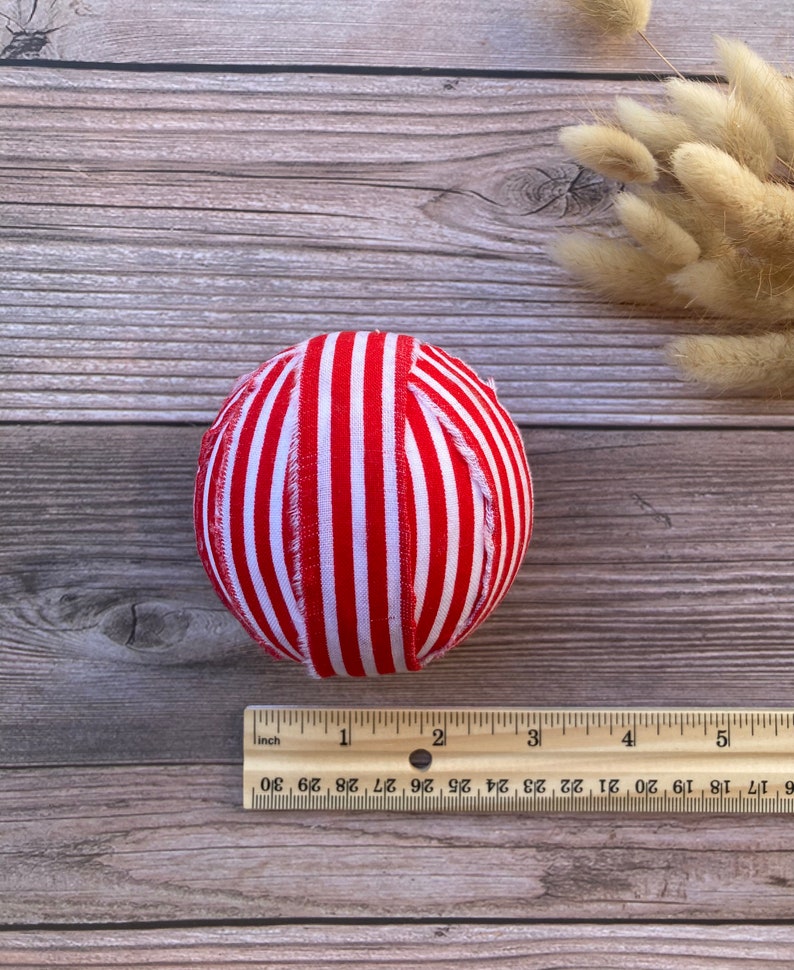 Puede incluir: Una bola de adorno de tela a rayas rojas y blancas. La bola tiene un di&aacute;metro de aproximadamente 5 cm.