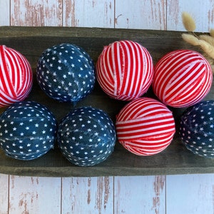 Puede incluir: Ocho bolas de tela en un cuenco de madera. Cuatro bolas son de rayas rojas y blancas y cuatro bolas son azules con estrellas blancas.