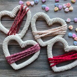 May include: Three white rope hearts with different colored yarn woven through them. The hearts are on a wooden background.