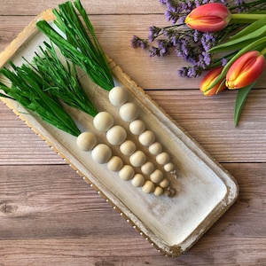 May include: Three wooden bead carrots with green paper grass stems on a white tray with a gold rim. The tray is on a wooden surface.