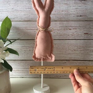 May include: A pink fabric bunny rabbit with a brown twine bow around its neck. The bunny is on a white wooden stand.