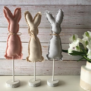 May include: Three fabric bunny figurines on white stands. The bunnies are pink, beige, and gray with a polka dot pattern. The bunnies are on a white wooden surface.