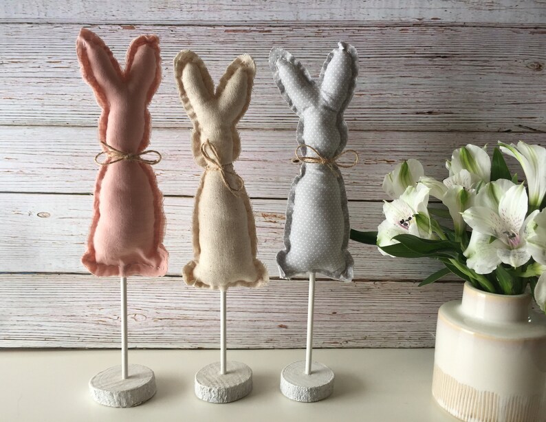 May include: Three fabric bunny figurines on white stands. The bunnies are pink, beige, and gray with a polka dot pattern. The bunnies are on a white wooden surface.