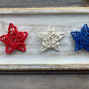 May include: Three woven star decorations in red, white, and blue. The stars are arranged in a row on a white wooden tray.