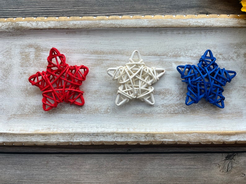 May include: Three woven star decorations in red, white, and blue. The stars are arranged in a row on a white wooden tray.