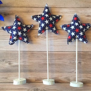 May include: Three red and white star-shaped fabric decorations on white sticks with white bases. The stars are on a wood background.