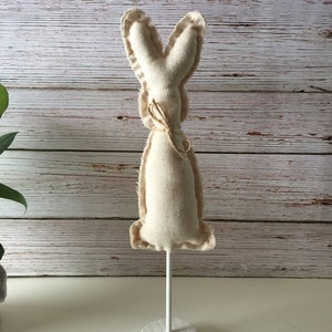 May include: A white fabric bunny rabbit with a brown twine bow, standing on a white wooden base.