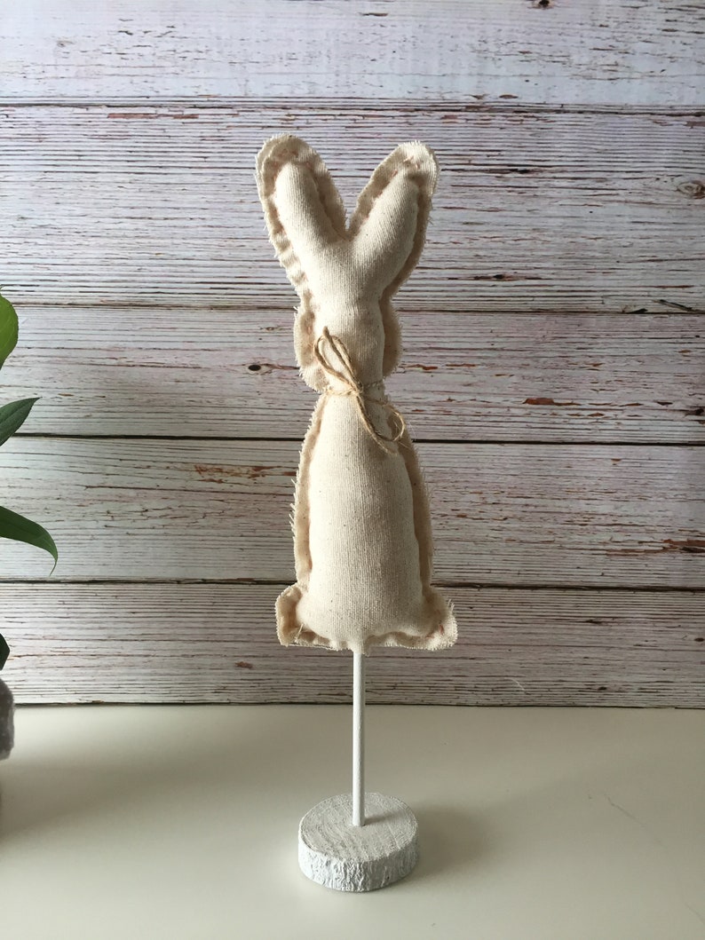 May include: A white fabric bunny rabbit with a brown twine bow, standing on a white wooden base.