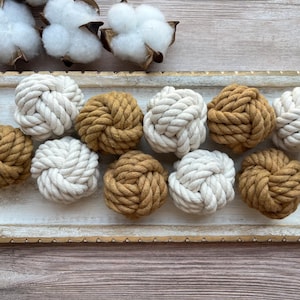 May include: A rectangular white tray with distressed gold edges holds ten spherical rope knots arranged in two rows. The knots alternate between solid off-white and mustard yellow colours, each featuring a complex, interwoven monkey fist design.