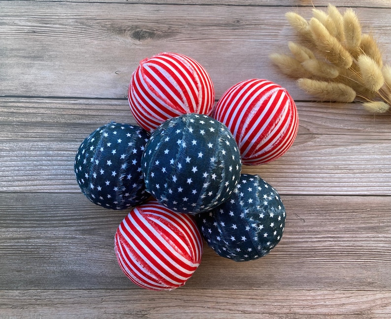 Puede incluir: Seis adornos de tela en rojo, blanco y azul con un patr&oacute;n de estrellas. Los adornos est&aacute;n dispuestos en c&iacute;rculo sobre una superficie de madera.
