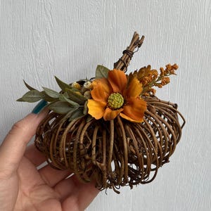 May include: A decorative pumpkin crafted from brown intertwined twigs, featuring a central orange flower with a green centre, accented by small green leaves and orange sprigs. The pumpkin has a twig stem wrapped with thin dark wire.