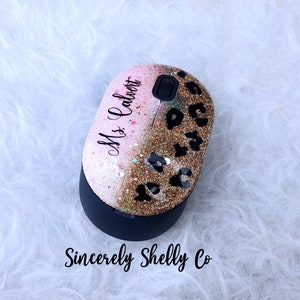 May include: A wireless computer mouse featuring a glittery finish that transitions from light pink to gold. The top surface displays a black leopard print pattern and the text Ms. Calvert in a script font. The base is matte black with a scroll wheel.