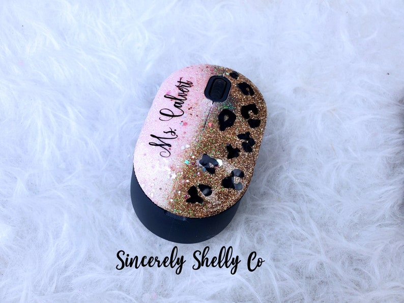 May include: A wireless computer mouse featuring a glittery finish that transitions from light pink to gold. The top surface displays a black leopard print pattern and the text Ms. Calvert in a script font. The base is matte black with a scroll wheel.
