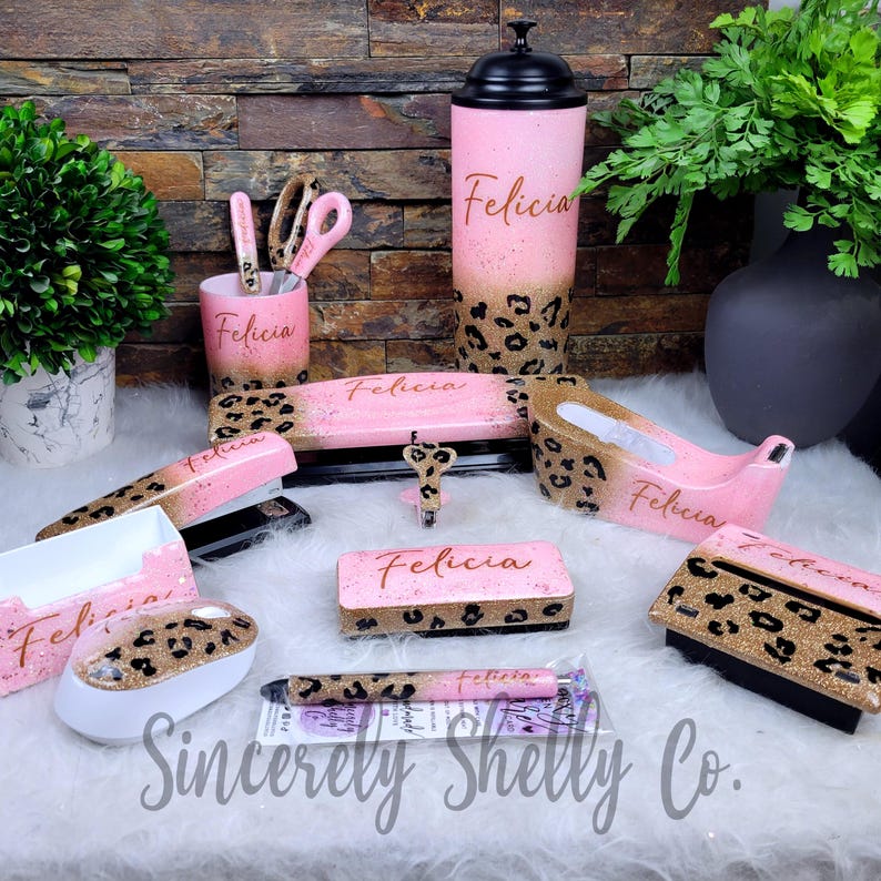 May include: A desk accessory set featuring a pink and gold glitter leopard print pattern. Items include a tumbler, pencil cup, stapler, tape dispenser, pen, mouse, and note holders, each personalized with the name Felicia in cursive script.