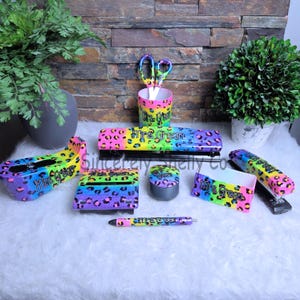 May include: A desk accessory set featuring a rainbow leopard print pattern with the name Mrs. Green printed on each item. The set includes a tape dispenser, a pencil cup with scissors, a hole punch, a pen, a stapler, and two small containers.