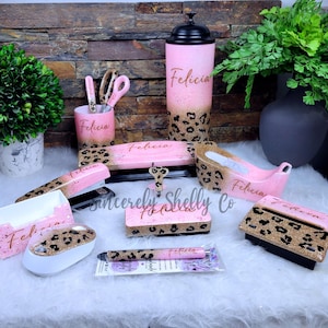 May include: A desk accessory set featuring a pink and gold glitter leopard print pattern with the name Felicia printed on each item. The collection includes a tumbler, pencil cup, stapler, tape dispenser, pen, mouse, and various office organizers.