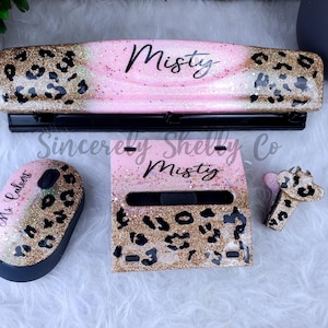 May include: A desk accessory set featuring a three-hole punch, a wireless computer mouse, a sticky note holder, and a stapler. Each item has a pink and gold glitter finish with a black leopard print pattern. The punch and holder are personalized with the name Misty, while the mouse features the name Ms. Calvert.