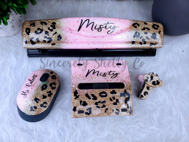 May include: A desk accessory set featuring a three-hole punch, a wireless computer mouse, a sticky note holder, and a stapler. Each item has a pink and gold glitter finish with a black leopard print pattern. The punch and holder are personalized with the name Misty, while the mouse features the name Ms. Calvert.