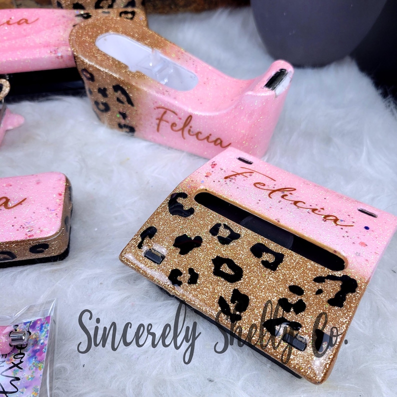 May include: A pink and gold glitter resin tape dispenser and a matching stapler, both featuring a black leopard print pattern and the name Felicia in cursive script. The items have a glossy finish and are designed for office desk use.