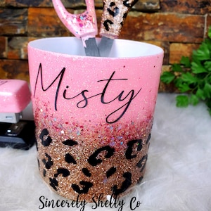 May include: A cylindrical desk organizer with a pink and rose gold glitter gradient finish, featuring a black leopard print pattern at the base and the name Misty in black script. A pink stapler and scissors are visible inside the container.