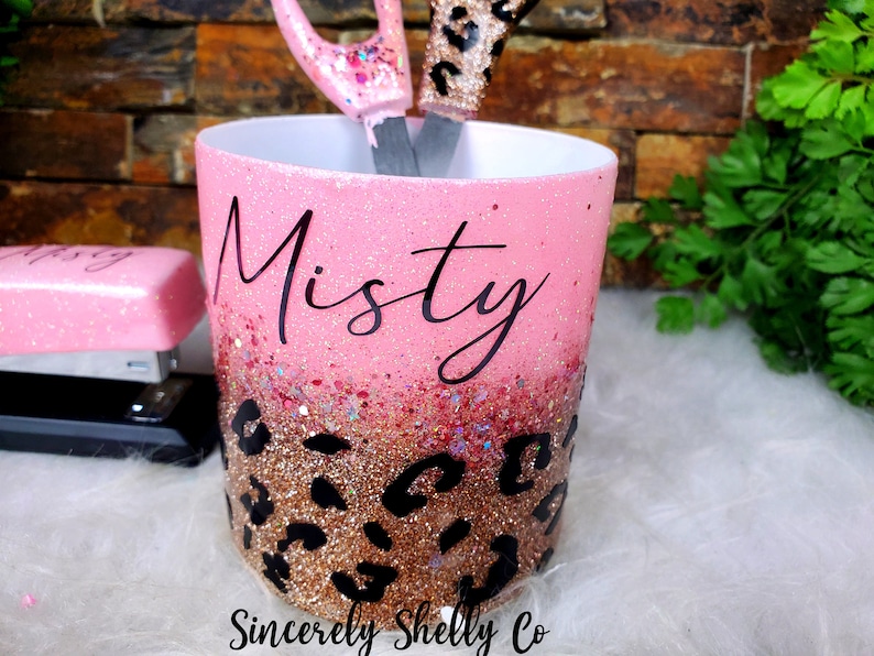May include: A cylindrical desk organizer with a pink and rose gold glitter gradient finish, featuring a black leopard print pattern at the base and the name Misty in black script. A pink stapler and scissors are visible inside the container.