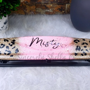 May include: A black and gold leopard print paper punch with the name "Misty" written in white cursive on a pink background. The punch has a black handle and a clear plastic top.