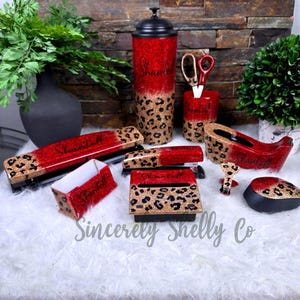 May include: A desk accessory set featuring a tumbler, pen holder with scissors, tape dispenser, stapler, note holder, and computer mouse. Each item has a red and gold glitter finish with a black leopard print pattern and the name Shantell.