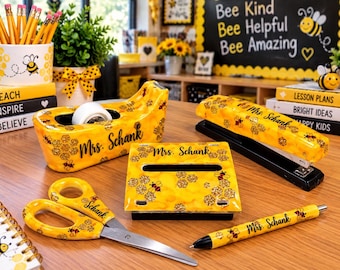 May include: A yellow desk accessory set featuring a bee and honeycomb pattern with the name Mrs. Schank printed on each item. The set includes a tape dispenser, a stapler, a pen, scissors, and a rectangular holder, all with black accents.
