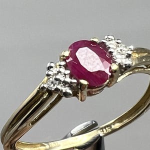 May include: A gold ring featuring a large oval ruby gemstone as the centerpiece. The ruby is a deep red color and is flanked by clusters of small, clear gemstones. The ring's band is a two-tone design, with gold and silver elements.