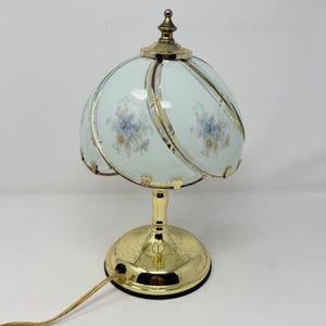 Pode incluir: Um candeeiro de mesa com base em metal dourado polido e cúpula de vidro em forma de domo. A cúpula apresenta um acabamento em azul pálido e branco com delicados padrões florais em azul e castanho, acentuados por nervuras de metal dourado.