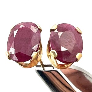 May include: A pair of ruby stud earrings. Each earring features a faceted, oval-shaped ruby gemstone set in a gold-colored metal. The rubies are a deep, rich red color, and the gold setting has a simple, classic design.