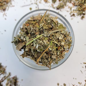 May include: A glass bowl filled with dried herbs, with additional dried herbs scattered around the bowl on a white surface.