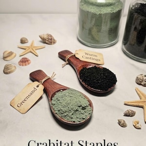 May include: Two wooden spoons filled with Greensand and Worm Castings, with attached tags. Jars of green and black powder are in the background. Seashells and starfish are scattered around the spoons. The text "Crabitats Staples" is at the bottom.