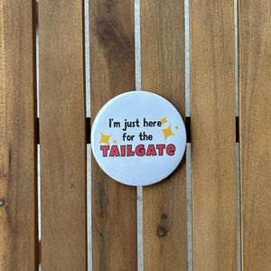 Puede incluir: Un botón blanco con el texto "I'm just here for the TAILGATE" en una fuente divertida, acentuado con estrellas amarillas. El botón está sobre un fondo de madera.