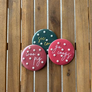 Puede incluir: Tres botones redondos y coloridos con texto blanco y detalles de puntos blancos. Uno es verde oscuro con el texto "sic em", otro es rojo con "woo pig" y el tercero es rojo con "go coogs".