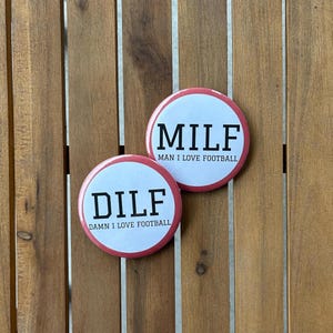 May include: Two round buttons with white centers and pink edges. One button reads "MILF MAN I LOVE FOOTBALL" in black text. The other button reads "DILF DAMN I LOVE FOOTBALL" in black text. The buttons are on a wooden surface.