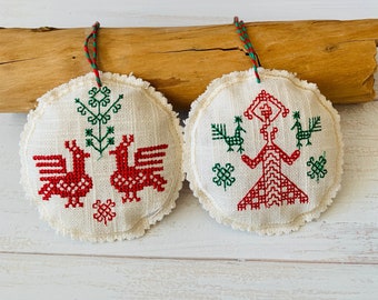 May include: Two white fabric ornaments with red and green embroidery. One ornament features two birds facing each other, while the other features a stylized figure with outstretched arms.