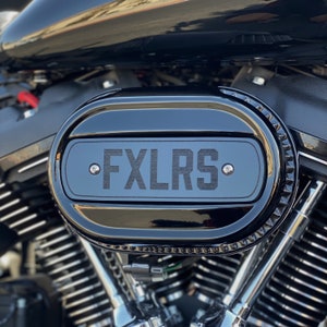 May include: Close-up of a black motorcycle air filter with a black oval plate that says "FXLRS" in white lettering.