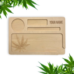 May include: A wooden rolling tray with a leaf design, a small circle, and a rectangular space for rolling. The tray has a slot for a grinder and a space for storing items. The tray is engraved with the text "YOUR NAME".