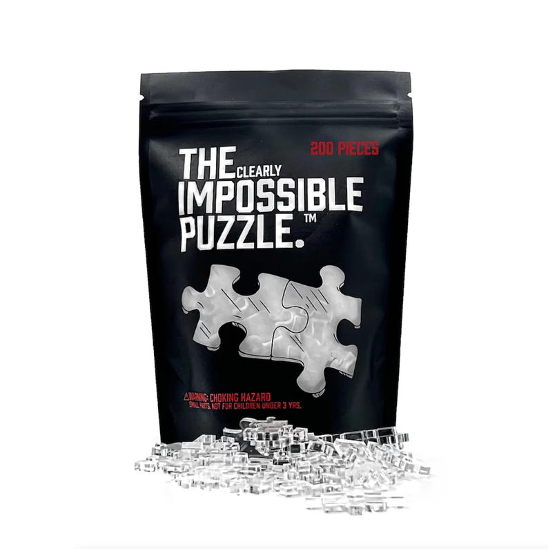 May include: A black bag with a white zipper containing a 200 piece clear plastic jigsaw puzzle. The bag has the text "The Clearly Impossible Puzzle" printed on it.