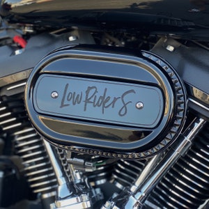 May include: Black motorcycle air filter with a chrome trim and a black oval plate with the text "Low Riders" in white lettering.