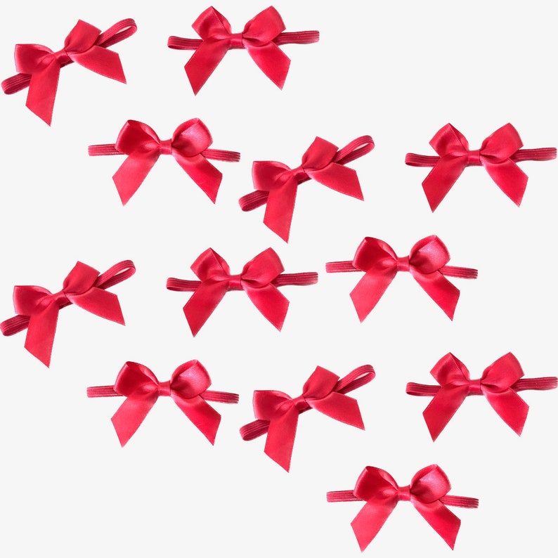 Puede incluir: Doce lazos de cinta de sat&eacute;n rojo con bandas el&aacute;sticas. Los lazos est&aacute;n dispuestos en un patr&oacute;n disperso sobre un fondo blanco.