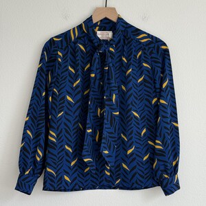 May include: A long-sleeve blouse in a deep blue color featuring a repeating black and yellow leaf-like pattern. The garment includes a high neck with a tie-front detail and buttoned cuffs. A label inside the collar reads Country Sophisticate Petite.