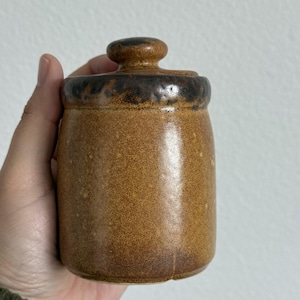 Peut inclure: Une tasse en céramique blanche avec une anse noire et le texte "But First, Coffee" en gras noir. La tasse se détache sur un fond flou, soulignant son design épuré.