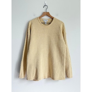 May include: A pale yellow, long-sleeved jumper with a textured, woven pattern. The jumper has a crew neck and is hanging on a wooden hanger against a white wall. The hem and cuffs are ribbed.