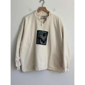 Op de afbeelding: Crèmekleurige sweatshirt met een hoge kraag en een vierkante, donkergroene afbeelding op de voorkant. De afbeelding lijkt een botanisch ontwerp te zijn. De sweatshirt heeft lange mouwen en een trekkoord in de hals. Het kledingstuk hangt aan een houten hanger.