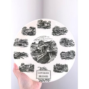May include: White ceramic plate featuring black and white illustrations of covered bridges. The plate has a central image and several smaller images around the edge. The words "COVERED BRIDGES" are printed on the plate.